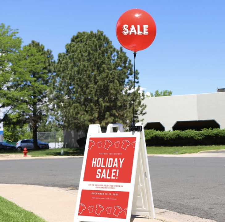 Red &ldquo;SALE&rdquo; balloon attached to a sidewalk A-frame sign advertising a holiday sale outside a storefront on a sunny day.